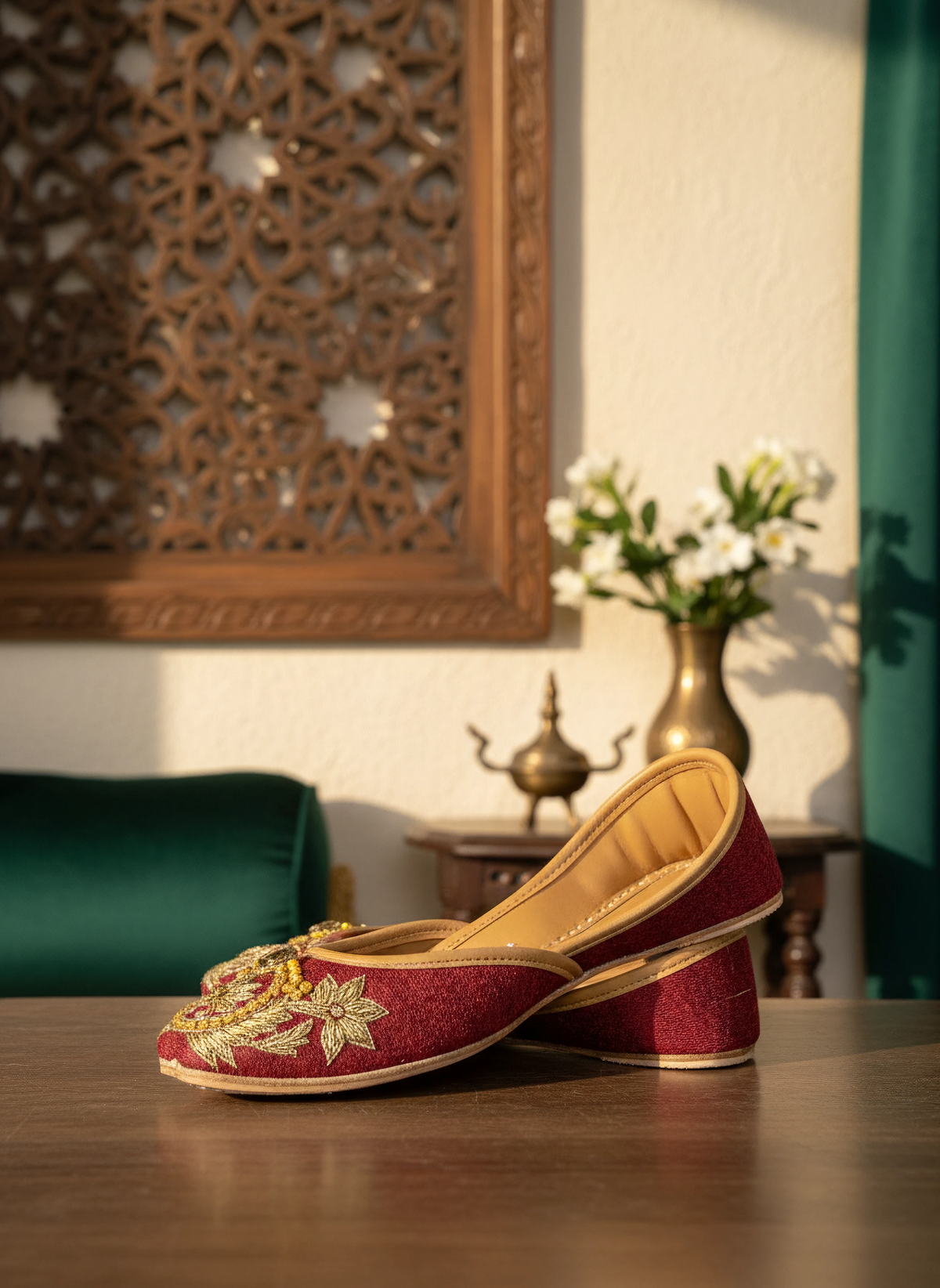 A pair of red embroidered flat shoes with gold detailing, styled on a light surface with a dark wooden carved screen, a green pillow, and a brass teapot in the background.