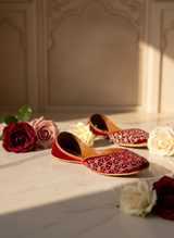 A pair of red traditional embroidered flat shoes with white beadwork, styled on a marble surface with red and white roses under warm sunlight.