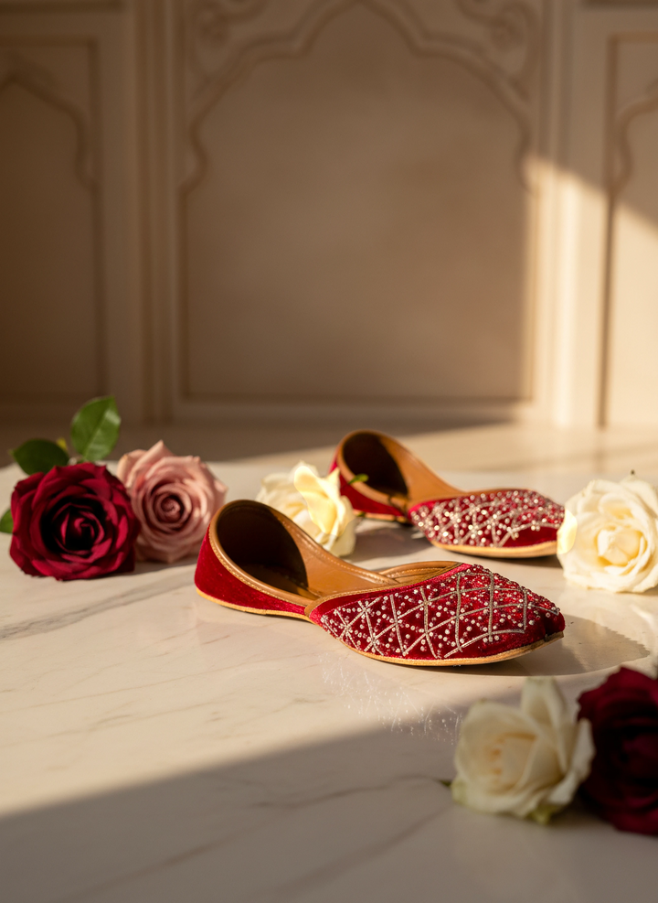 A pair of red traditional embroidered flat shoes with white beadwork, styled on a marble surface with red and white roses under warm sunlight.